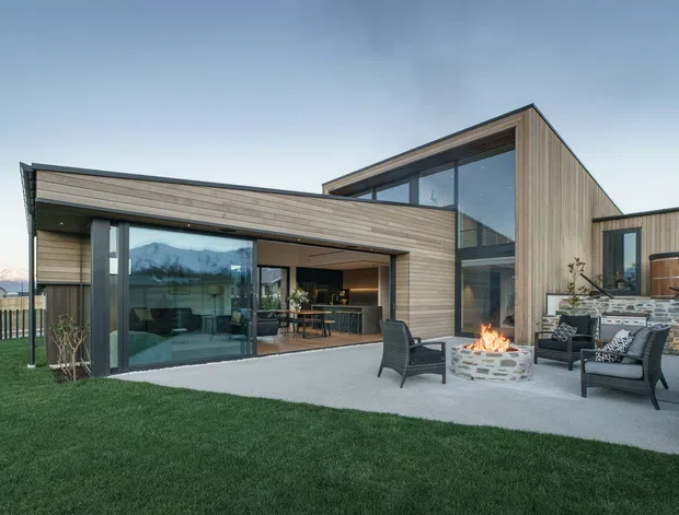 Contemporary single-storey residential house viewed from the landscaped outdoor area, with intersecting low-pitched roof forms, extensive floor-to-ceiling glazing, and vertical timber-clad walls. A covered patio and outdoor lounge surround a circular open fire pit, while the interior kitchen/dining area is visible through the glazing. Stone paving and retaining elements connect the lawn to the house.