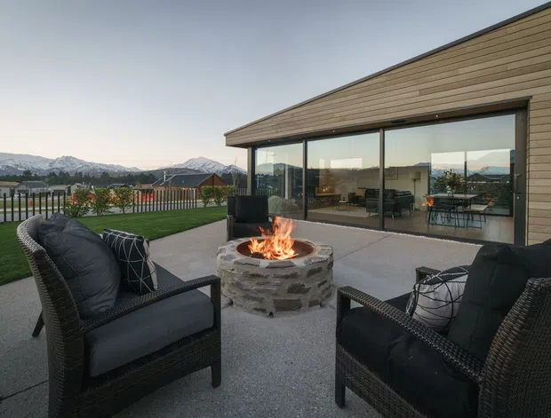 Contemporary single-storey home with a long, low-pitched roofline and horizontal timber cladding, opening onto a broad paved terrace. Dark woven outdoor armchairs surround a round stone outdoor fire pit, while extensive glazing reveals a bright interior dining and living area. A lawn, fenced property edge, and distant mountain range form the background.