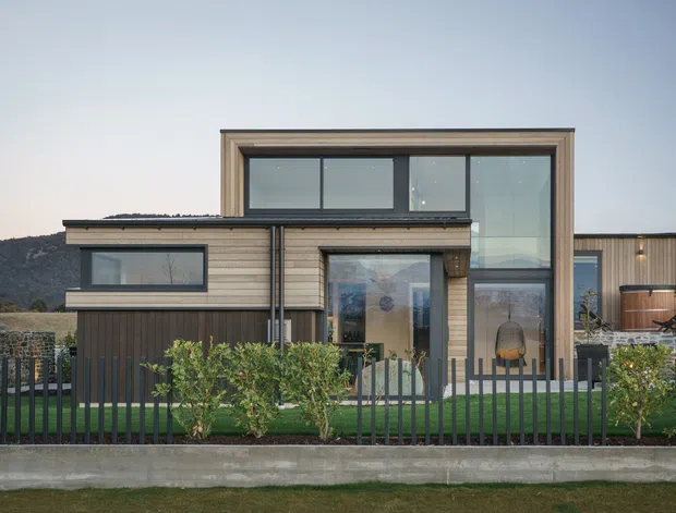 Contemporary multi-level residential facade with a strong rectangular composition, extensive dark-framed glazing, horizontal timber-look cladding, and a recessed central entry. A dark metal fence and landscaped planting sit in the foreground, with a low mountain or hillside visible behind the property.