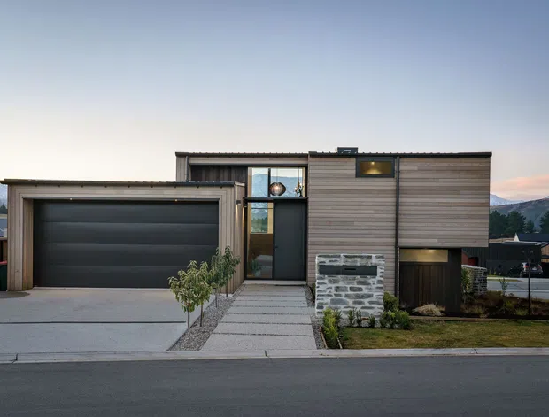 Front exterior view of a contemporary multi-level residence with a prominent attached single-car garage, horizontal timber-clad upper facade, dark metal-framed glazing, and a stone-clad entry feature wall. The property sits on a suburban street with mountains visible in the distance.