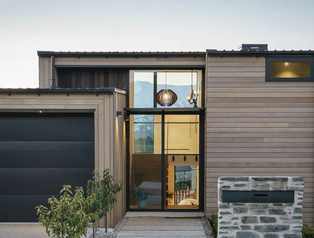 Front exterior of a contemporary single-storey residence with horizontal timber cladding, a dark sectional garage door, tall glazed entry, flat rooflines, and a masonry feature wall beside a concrete path. The facade has a restrained neutral palette and strong vertical entry glazing.