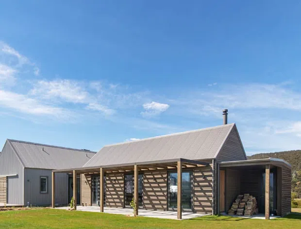 Single-storey rural home with a simple gabled metal roof, grey corrugated exterior cladding, and a long timber-framed covered outdoor area. The building sits in an open grassy setting with hills behind it under a clear blue sky. A timber privacy screen and stacked firewood are visible beneath the covered zone, with large glazed openings connecting the interior to the exterior.