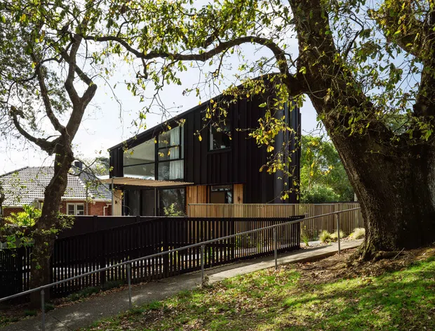 Exterior view of a contemporary two-storey dark-clad house set among mature trees, with large glazed openings, a timber balcony or terrace, and an accessible sloping walkway with metal handrails. A neighbouring traditional tiled-roof house is partially visible behind it.