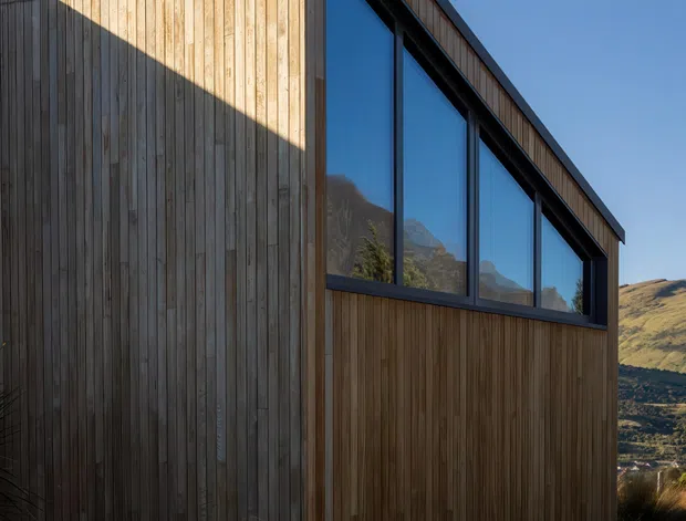 Exterior view of a compact contemporary timber-clad building with a steeply sloping roofline, dark metal window frames, expansive glazing, and a chimney. The vertical natural timber cladding is prominent, with mountainous terrain reflected in the windows and visible beyond the building.