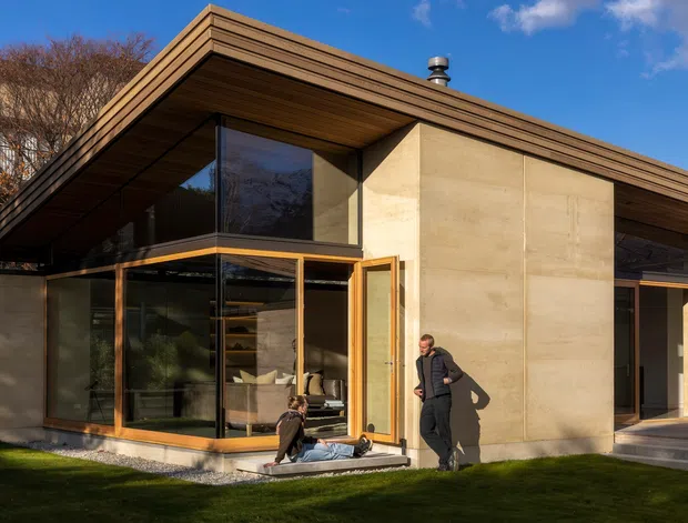 Exterior view of a low-slung contemporary house with broad projecting roof planes, warm timber soffits and fascia, expansive floor-to-ceiling glazing, timber-framed doors and windows, and large smooth concrete wall panels. The corner glazing reveals a furnished living area inside, while people and lawn occupy the foreground.