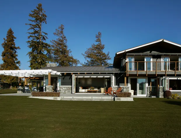 Exterior view of a large two-storey suburban residence set behind an expansive lawn. The home has a dark pitched gable roof, light exterior walls, extensive glazing, a prominent upper-level balcony with dark timber-toned railings, and a covered outdoor entertaining area/pergola extending to the left. Mature trees and landscaped garden elements frame the property.