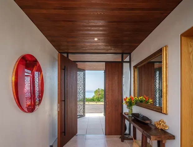 A narrow, elegant residential entry hall with a richly stained timber-lined ceiling, pale plaster walls, and a large patterned runner over pale tiled flooring. Dark timber doors open toward a bright exterior terrace, with decorative patterned screens visible beyond. A slim timber console table, ornate gold-framed mirror, red wall artwork, and decorative objects add warmth and character.