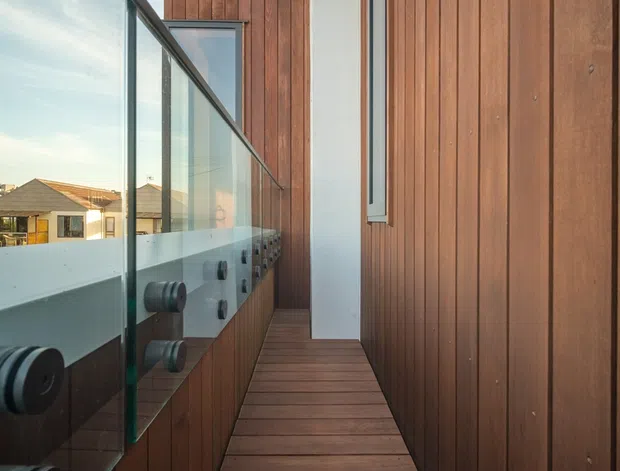 Narrow elevated exterior walkway or balcony beside a modern building, with dark timber decking and vertical timber wall cladding. A clear glass balustrade with dark metal fittings lines the open side, overlooking neighbouring rooftops. The composition emphasises the linear passage, warm wood surfaces, and clean contemporary detailing.