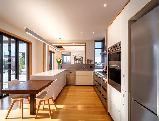 Bright, open-plan contemporary kitchen with extensive white built-in cabinetry, integrated stainless-steel appliances, a long central island, and warm timber flooring. A compact timber dining table with white chairs sits beside full-height glazed doors, while timber-framed openings, exposed timber ceiling detailing, and generous daylight create a warm, refined interior.