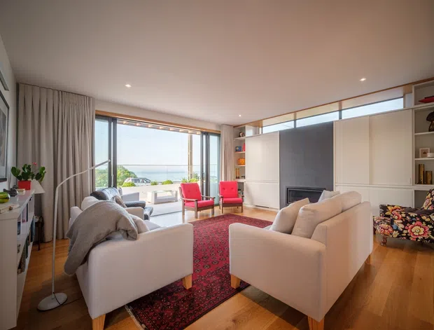 Bright, warmly furnished living room with polished timber flooring, two cream armchairs and a cream sofa arranged around a red patterned rug and a built-in fireplace. Full-height glazing and sliding doors open toward a balcony or terrace with a distant coastal outlook. Built-in shelving and cabinetry line the fireplace wall, while curtains, artwork, plants, and colourful occasional chairs add character.