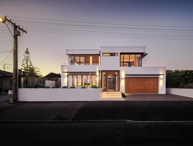 Twilight street-facing photograph of a contemporary two-storey residence with a crisp white rendered facade, broad horizontal forms, large glazed openings, warm interior lighting, a recessed central entry, and an integrated double garage with a timber-toned door. The home sits behind a low white boundary wall on an urban suburban street, with neighbouring houses and overhead utility wires visible.