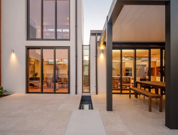A dusk view of a contemporary residential courtyard between two modern building volumes. Large glazed doors and windows reveal warm-lit living and dining areas, while a covered outdoor dining space with timber furniture occupies the right side. The courtyard is paved in large pale stone-look slabs and incorporates narrow linear water features or reflecting channels. Dark metal framing, flat rooflines, minimalist detailing, and warm exterior wall lights define the composition.