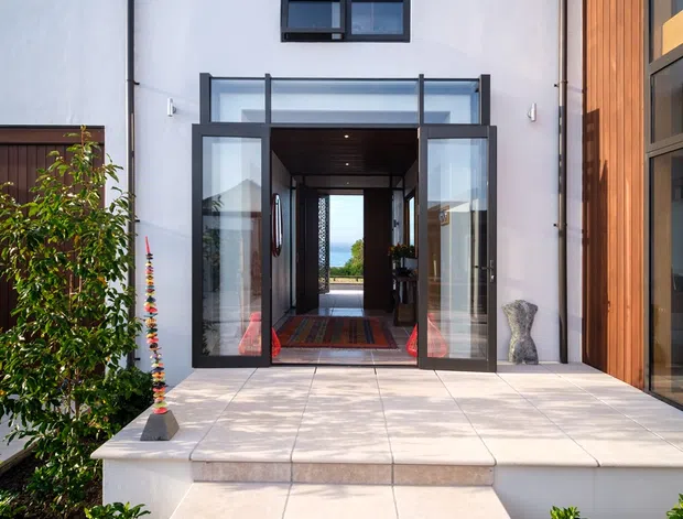 Exterior view of a contemporary residential entrance. Broad pale tiled steps lead to a raised terrace and fully glazed double entry doors, with a dark-framed transom and sidelights. The facade combines smooth white render with a vertical warm timber-clad wall, tall black exterior sconces, and simple landscaped planting. A hallway is visible through the open doors, aligned toward a distant blue view.