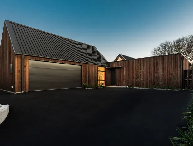 A contemporary residential exterior featuring a detached-looking garage or outbuilding with a steep gabled roof, dark standing-seam metal roofing, vertical timber cladding, and a large concealed-panel garage door. A matching timber-clad boundary wall and gated entry enclose a broad dark paved driveway, with subtle planting along the edges.