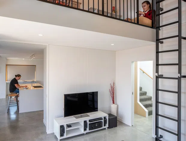 Bright contemporary double-height living room with polished concrete flooring, white walls and built-in cabinetry, a black metal mezzanine balustrade and ladder, and a timber-lined ceiling. A compact kitchen is visible to the left, with a stairway leading upward at the rear.