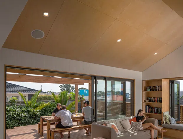 Bright contemporary open-plan living room with a large timber-lined sloping ceiling, polished concrete floor, extensive sliding glass doors, and direct connection to a timber deck. A grey sofa, patterned ottoman, rug, built-in timber shelving, and outdoor dining area are visible, with strong sunlight and garden views.