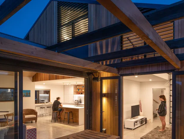 Twilight view of a contemporary multi-level timber-clad home arranged around an open entertaining deck and courtyard. Heavy exposed timber beams frame glazed openings into a bright kitchen and living area, with vertical timber wall cladding, dark structural steel elements, and a built-in timber dining table and benches on the deck.