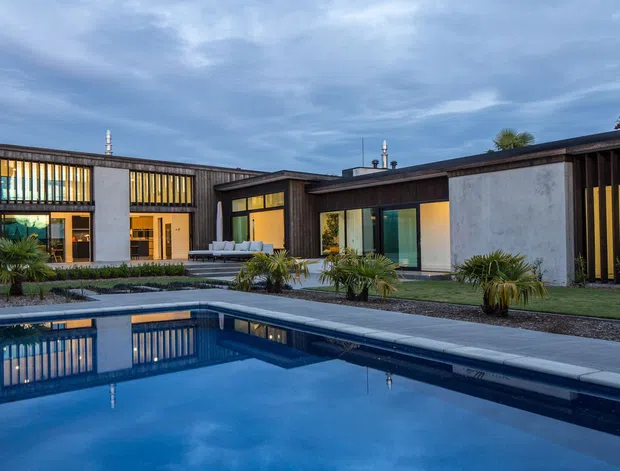 A low, modern single-storey residence arranged around a landscaped swimming pool. The building features dark vertical timber cladding, smooth light rendered walls, extensive warm-lit glazing with vertical fins, and broad flat rooflines. The pool and paved terrace dominate the foreground, with structured planting around the courtyard-like outdoor space.