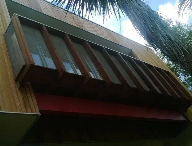 Upward-looking exterior detail of a contemporary timber-clad building, featuring vertical warm wood boards, a projecting window bay with repeated timber fins or exposed framing, and dark metal roof and fascia elements. Tropical foliage partially frames the facade.