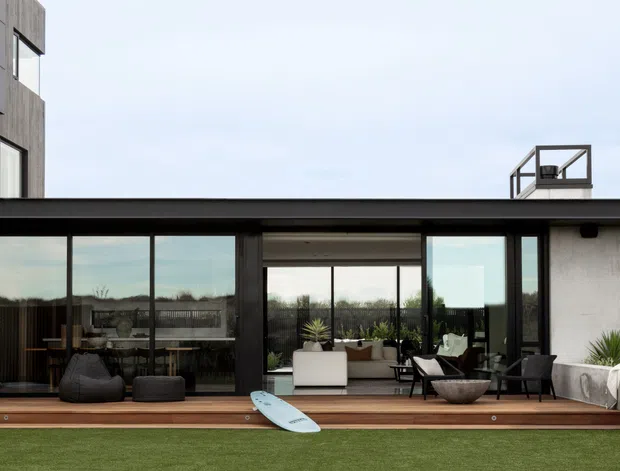 Contemporary flat-roof residential exterior viewed from a landscaped lawn, with extensive floor-to-ceiling glazing opening onto a timber deck. The dark-framed glass facade reveals a furnished open-plan living and dining area, while a white rendered side wall, rooftop structure, outdoor seating, and a surfboard contribute to the relaxed modern setting.