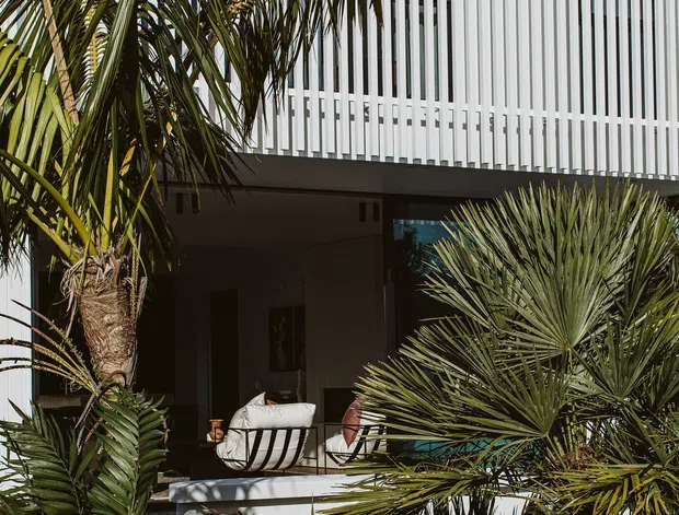 Exterior view of a contemporary multi-level building partially screened by lush tropical and subtropical planting. The facade features crisp white vertical slatted screening above a shaded ground-level terrace, with outdoor seating visible beneath the overhang.