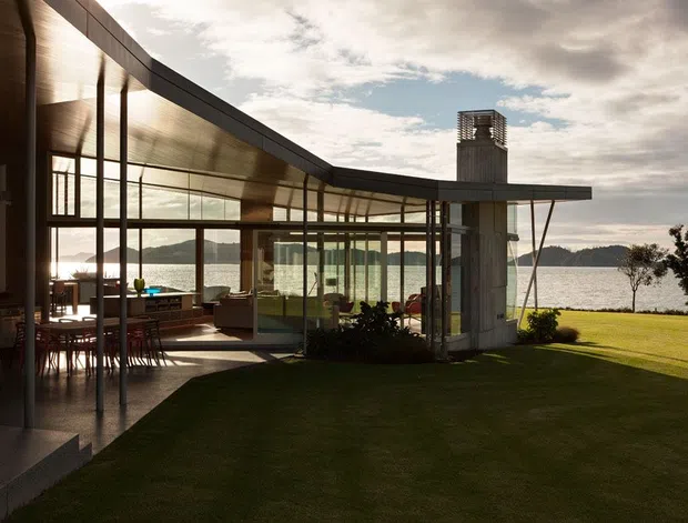 Exterior view of a low-slung modern waterfront residence with broad roof overhangs, extensive floor-to-ceiling glazing, dark metal framing, and warm timber soffits. The open-plan dining and living areas are visible through the glass, opening onto a manicured lawn beside the water. A sculptural chimney volume and slim angled supports reinforce the contemporary pavilion-like form.