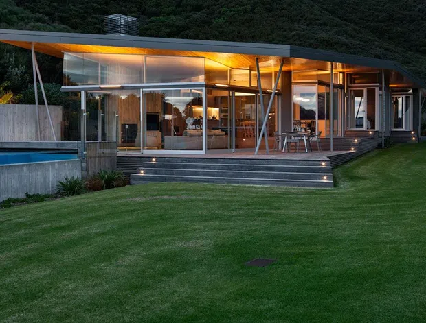 Exterior dusk view of a low, elongated modern house set against a densely vegetated hillside. The pavilion-like structure has extensive floor-to-ceiling glazing, slender structural posts, a broad shallow roof overhang, warm interior lighting, and a raised timber deck accessed by wide steps with integrated lights. A swimming pool and concrete retaining walls sit to the left, with manicured lawn in the foreground.