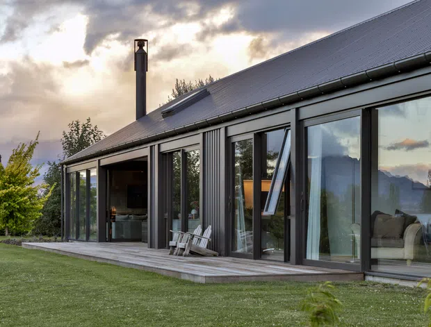 Exterior view of a contemporary single-storey home with a steep pitched metal roof, dark vertical metal cladding, extensive floor-to-ceiling glazing, and a timber deck opening onto a lawn. The glazed facade reveals a living area inside, while the surrounding landscape includes mature trees and distant mountainous terrain beneath a dramatic cloudy sky.