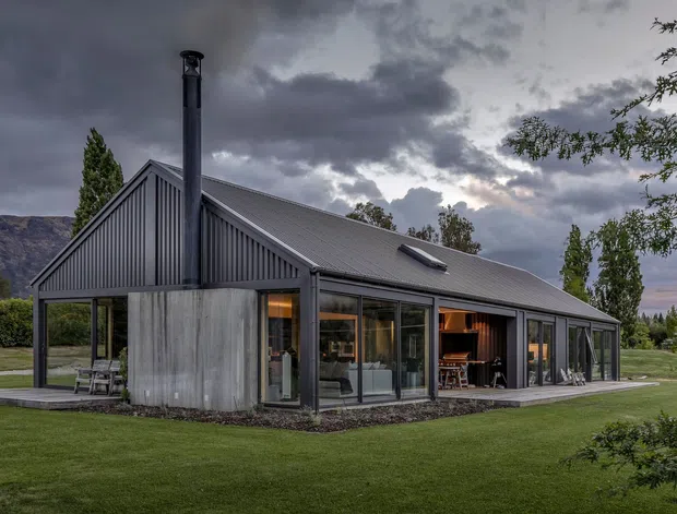 A modern single-storey gabled house set within a manicured lawn and landscaped grounds. The building has dark grey vertically ribbed metal cladding, a pitched metal roof with a skylight, extensive floor-to-ceiling glazing, and a tall external chimney. Warm interior lighting is visible through the windows and open doors, with mountains and dramatic cloud cover in the background.