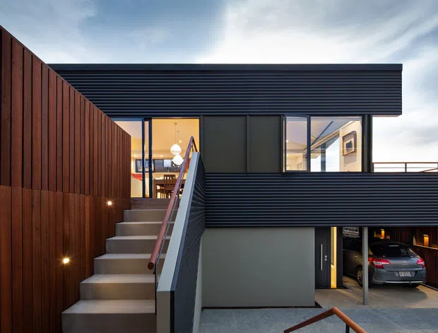 Exterior view of a contemporary multi-level residence with a flat roof, dark horizontally ribbed cladding, and warm vertical timber screening. An external concrete stair leads to an upper-level glazed living area, while an open garage with a parked car is visible below.