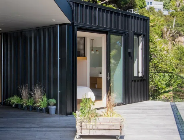 Exterior view of a compact contemporary dark-clad dwelling or extension opening onto a timber deck. The facade features vertically ribbed dark metal cladding, large glazed doors and windows, and a sheltered entry. A bedroom and part of a light-toned interior are visible through the open doorway. Planters and landscaped greenery frame the deck, with a hillside residential setting beyond.