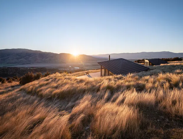 A low-profile contemporary building is partially nestled into a rolling, grass-covered hillside at sunset. The dark pitched roof and chimney emerge from tall golden tussock grass, with expansive mountainous terrain and a broad valley visible beyond. The scene has a secluded alpine or rural character and warm late-day light.