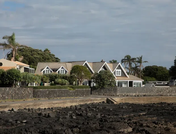 Exterior view of a large coastal-style residential house with multiple steep gables, grey shingle-like roof sections, white trim, and pale weatherboard walls. The property sits behind a dark stone boundary wall, with palm trees and rugged black volcanic rock in the foreground, suggesting a coastal or volcanic shoreline setting.