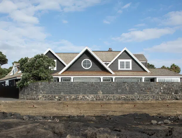 Exterior view of a large, low-rise residential house behind a dark volcanic-stone boundary wall. The home has multiple steep front-facing gables, grey shingle-like roof surfaces, white trim, dormer windows, and a circular feature window. It is set in a stark rocky landscape under a bright partly cloudy sky.