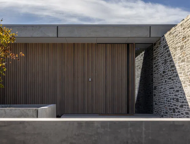 A minimalist contemporary building facade with a broad low-profile concrete roofline and parapet, a dark vertical timber-slatted entrance wall, and a contrasting rough masonry side wall. A recessed glazed or dark-framed opening sits between the timber and stone surfaces, with a concrete boundary wall in the foreground and a small deciduous tree at the edge.