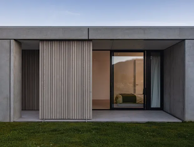 Front-facing exterior view of a minimalist single-storey dwelling with a flat roof and monolithic grey concrete facade. A recessed entry is screened by vertical timber battens, while a large dark-framed glazed opening reveals a simply furnished interior. The building sits directly beside a lawn under a clear sky.