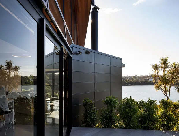 Exterior-facing view of a contemporary waterfront residence, showing dark metal-framed glazing, a timber-lined soffit, a projecting grey metal-clad volume, and a tall cylindrical chimney. A terrace and landscaped planting overlook the water and opposite shoreline.
