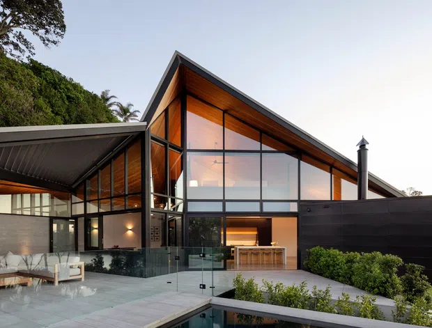 Exterior architectural photograph of a contemporary multi-level residence with a prominent steep gable roof, extensive floor-to-ceiling glazing, dark metal framing, and warm timber-lined soffits and interior surfaces. A covered outdoor entertaining area, paved terrace, landscaped planting, and a partially visible pool sit in front of the house. The design has a strong indoor-outdoor connection and a dramatic vaulted roof form.