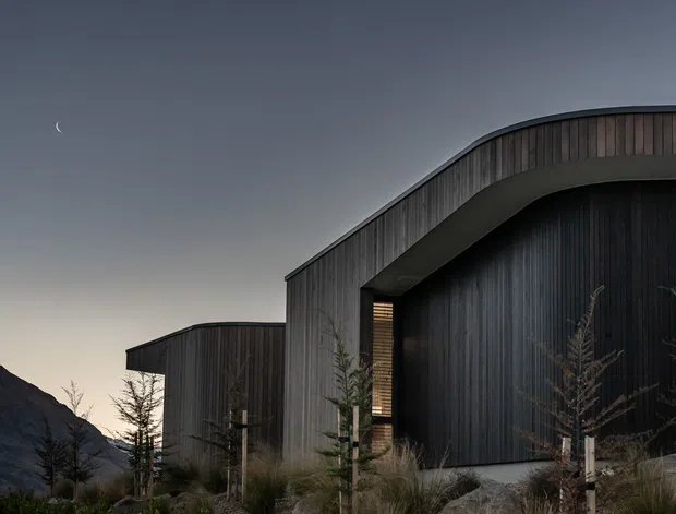 Exterior architectural photograph at dusk showing a contemporary dark-clad building with sweeping curved rooflines and vertical timber-like cladding. The structure sits against a mountainous landscape, with sparse landscaped planting in the foreground and a crescent moon visible in the sky.