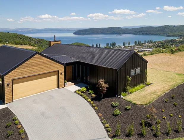 Aerial view of a contemporary single-storey hillside home with dark standing-seam metal roofing and dark vertical cladding, complemented by a warm timber-clad garage wing. The property has a broad concrete driveway, landscaped planting beds, and expansive views across a lake and surrounding rural hills.