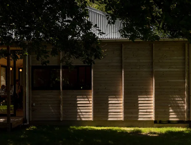 A shadowed exterior view of a low-rise contemporary building, with extensive horizontal timber weatherboard cladding, dark-framed glazing, and a partially visible covered outdoor seating area at left. Dense mature trees frame the facade, while a corrugated metal upper roof or screened volume rises behind it. Warm sunlight creates strong striped shadows across the timber walls.