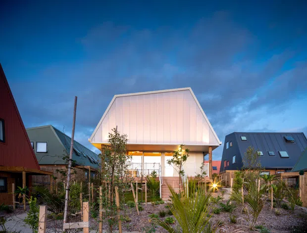 Exterior dusk view of a compact contemporary house in a landscaped residential development. The central home has a prominent steep gambrel-like metal-clad roof form, pale vertical wall cladding, an illuminated covered upper-level balcony or porch, and a planted gravel garden with young trees and palms. Neighbouring dark-clad houses are visible on both sides.