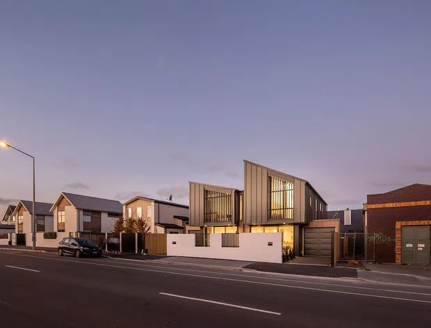 Twilight streetscape showing a row of contemporary residential buildings. The main subject is a narrow multi-level home with a sculptural pitched roof, vertical timber-toned cladding, large glazed openings, a recessed entry, garage, and a low white boundary wall. Adjacent modern homes and a dark brick building line the broad paved street.