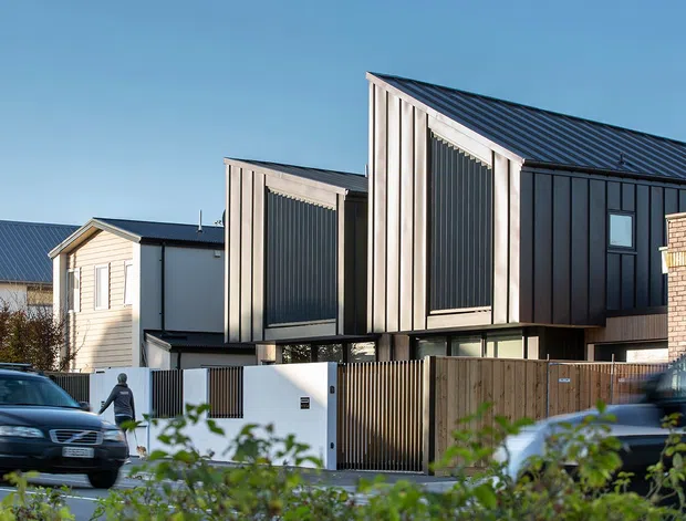 Street-facing view of a contemporary multi-level residential building with a dramatic steeply angled dark metal roof, black vertical metal cladding, pale timber-toned vertical facade panels, and elevated upper volumes above a secure timber and metal fence. Neighbouring homes and parked vehicles are visible in the foreground and background.