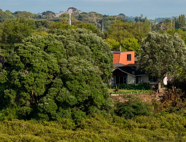 A telephoto exterior landscape view across calm water toward a small dark-clad house nestled among dense mature greenery. The house has a prominent bright orange-red upper roof or cladding element, while utility poles and distant tree-covered land sit behind it beneath a broad pale blue sky.