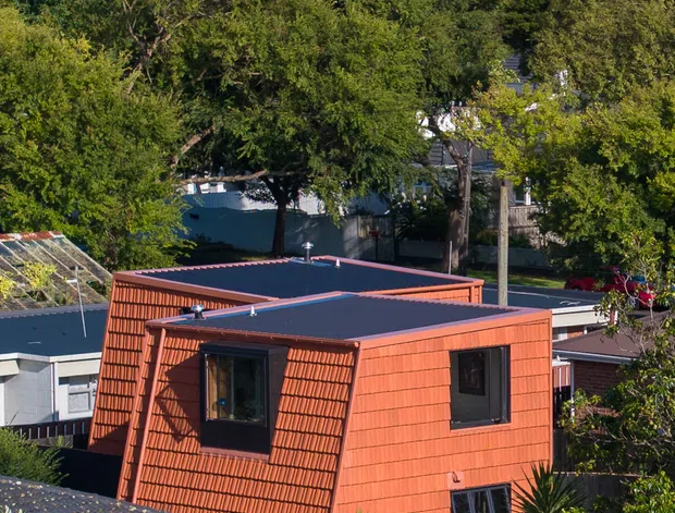 Elevated exterior view of a distinctive compact residential building in a suburban, tree-filled setting. The house has a strongly faceted, tapered form with vivid terracotta-orange textured cladding, dark flat roof planes, dark-framed windows, and surrounding established vegetation. Other low-rise homes, utility lines, and telecommunications structures are visible in the background.
