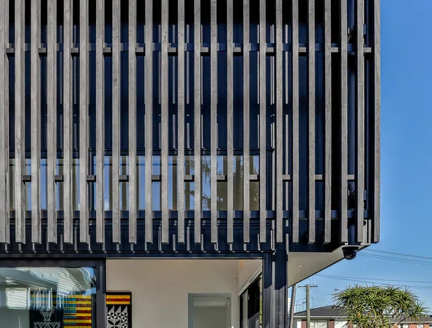 Front exterior view of a contemporary multi-storey building with a strong vertical facade screen made from closely spaced dark timber or metal fins. The upper level projects slightly over a glazed ground-floor entry and living area, creating a pronounced cantilevered form. The facade combines dark framing, extensive glazing, and rhythmic vertical shading elements. A paved path, lawn, hedge, and neighbouring suburban houses are visible around the entrance.