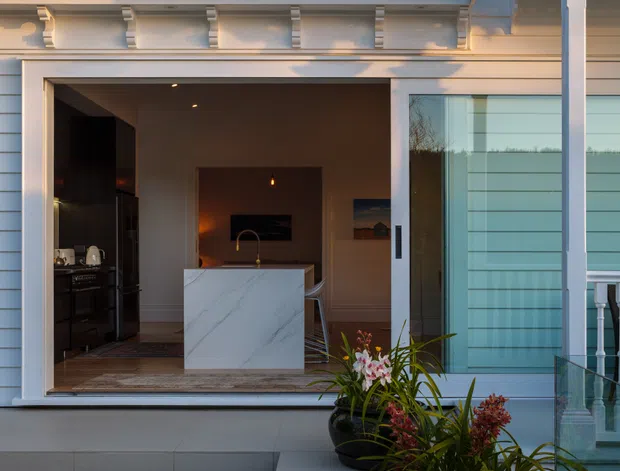 A refined exterior view of a heritage-style white residence opening onto a contemporary kitchen. The facade features weatherboard cladding, ornate painted timber fretwork, decorative columns and elaborate cornice detailing. Broad tiled steps lead to a large glazed opening, revealing a dark kitchen with a prominent white veined stone island. Potted plants and a glass balustrade frame the foreground.